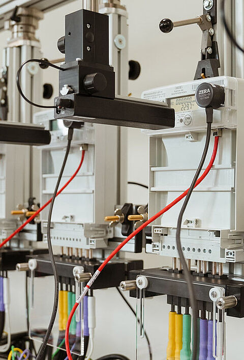 Reihe von Stromzählern in einer Prüfumgebung, verbunden mit farbigen Kabeln und mechanischen Einstellarmen zur Kalibrierung und Genauigkeitsprüfung – typische Szene aus einem Labor für Elektrotechnik oder Qualitätssicherung.