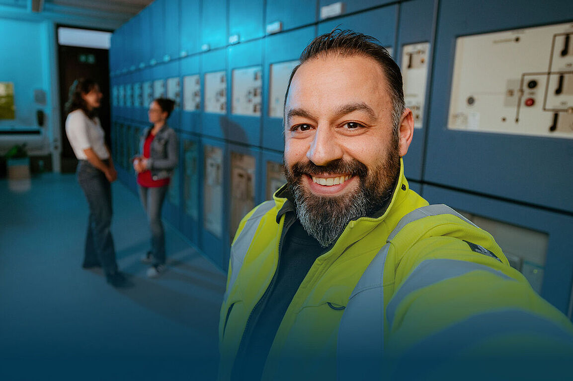 Ein lächelnder Techniker in einer gelben Warnjacke macht ein Selfie vor einer Reihe von elektrischen Schalttafeln in einem Technikraum. Im Hintergrund unterhalten sich zwei Kolleginnen.
