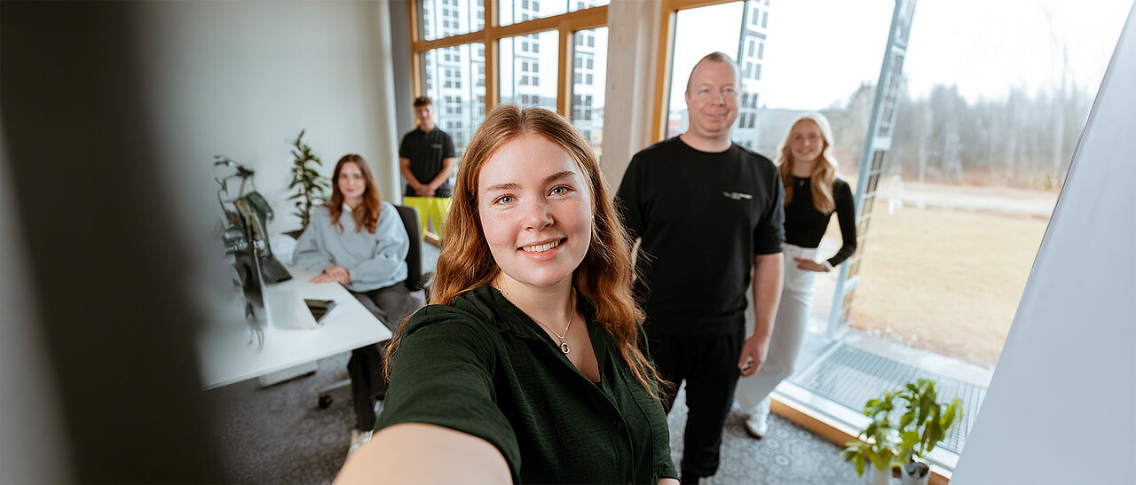 Mehrere Personen in einem hellen Büro mit Schreibtisch, Fenstern und Pflanzen.