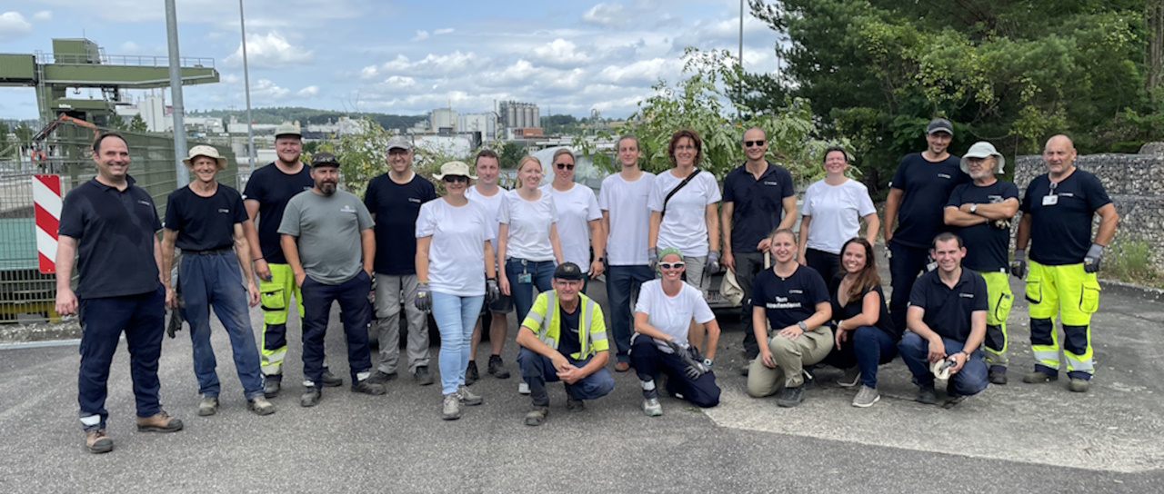 Eine Gruppe von etwa 30 Mitarbeitenden der naturenergie Gruppe steht im Freien auf einer gepflasterten Fläche. Im Hintergrund sind Grünflächen und industrielle Strukturen zu sehen. Die Personen tragen verschiedene Arbeitskleidungen, darunter Warnwesten, Sicherheitshelme und Freizeitkleidung. Ihre Gesichter sind aus Datenschutzgründen unkenntlich gemacht. Die Mitarbeitenden beteiligen sich an einer Aktion zur Bekämpfung invasiver Neophyten am Wasserkraftwerk Rheinfelden, um die heimische Flora und Fauna zu schützen.